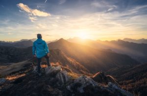 Homem sobre montanha, olhando o pôr do sol ao horizonte. Imagem simboliza sucesso e superação de desafios.