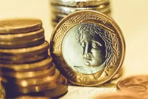 Moeda de real em pé, encostada em pilha de moedas deitadas.