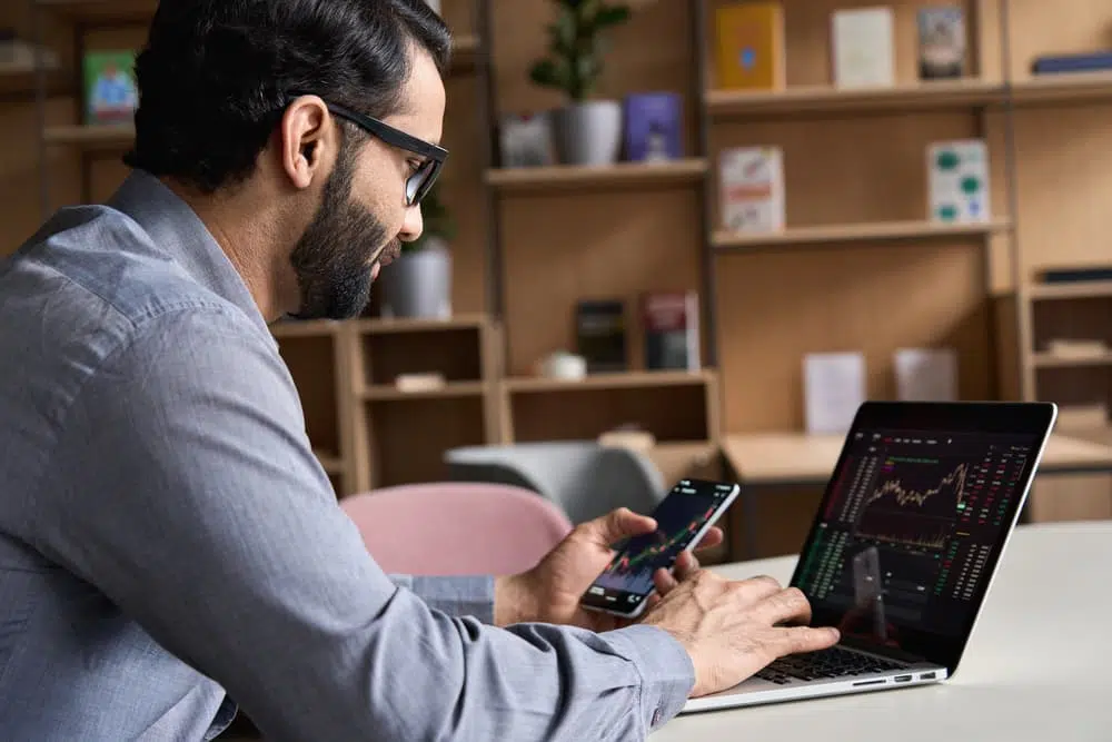 Homem vendo gráficos de investimentos em tela de celular e de computador.