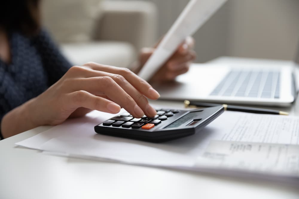 Mulher digitando em calculadora, com papéis e computador ao fundo, simbolizando o trabalho de contabilidade.