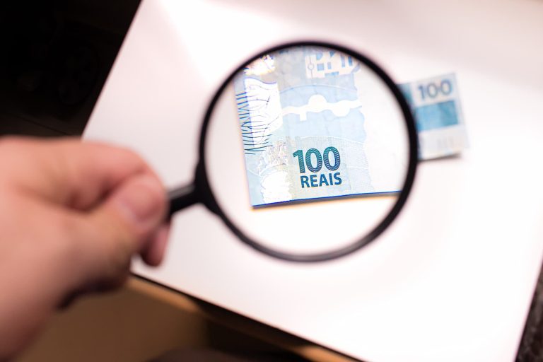 imagem mostra uma lupa direcionada para uma nota de cem reais.