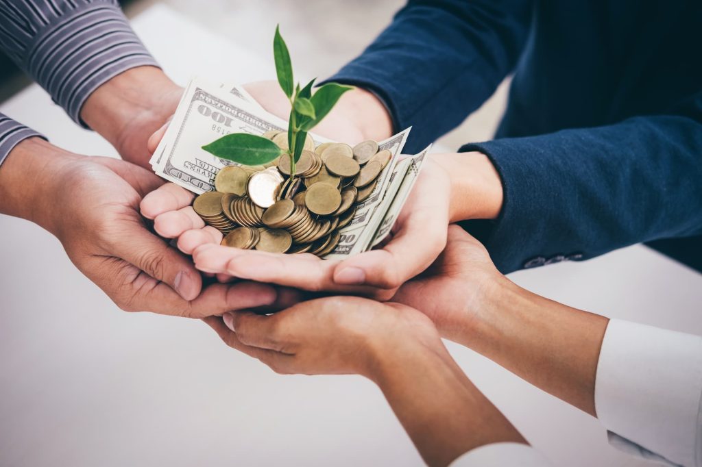 imagem mostra mãos de pessoas unidas com notas e moedas de dinheiro e uma planta que faz referência ao crescimento do patrimônio.